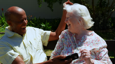 Front-view-of-active-mixed-race-senior-couple-using-mobile-phone-in-the-garden-of-nursing-home-4k
