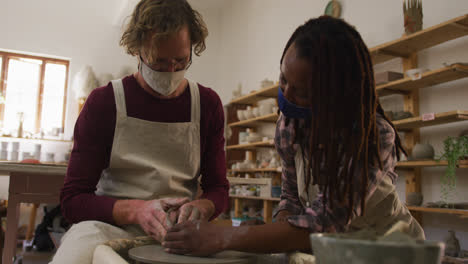 Diverse-male-and-female-potters-wearing-face-masks-creating-pottery-on-potters-wheel-at-pottery-stud
