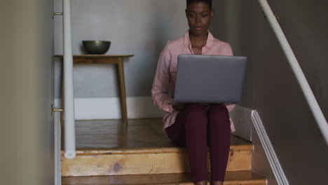 African-american-woman-using-laptop-while-sitting-on-stairs-working-from-home