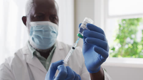 Male-african-american-doctor-wearing-face-mask-pulling-out-covid-19-vaccine-into-syringe