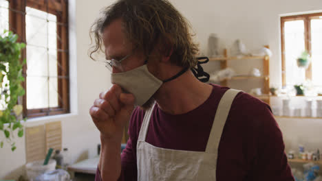 Male-caucasian-potter-wearing-face-mask-and-apron-coughing-at-pottery-studio