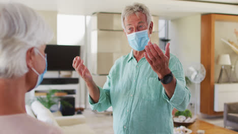 Senior-caucasian-couple-wearing-face-masks-greeting-each-other-by-touching-elbows-at-home
