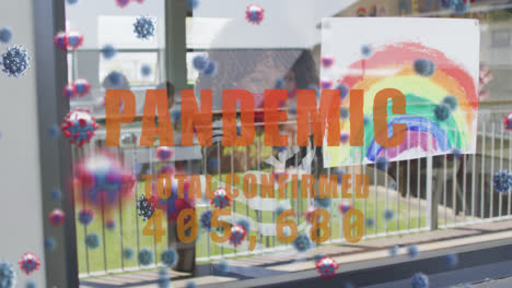 Pandemic-text-with-increasing-cases-against-boy-holding-rainbow-painting-against-window