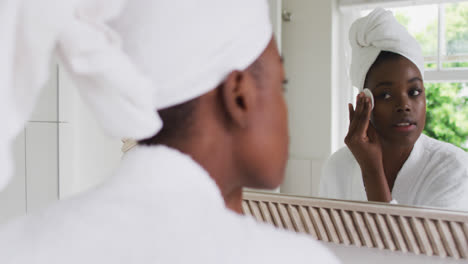 Afroamerikanische-Frau-Im-Bademantel-Reinigt-Ihre-Haut-Mit-Einem-Wattepad,-Während-Sie-In-Den-Spiegel-Schaut