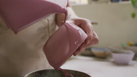 Close-up-view-of-female-potter-pouring-paint-from-a-jug-on-pot-at-pottery-studio