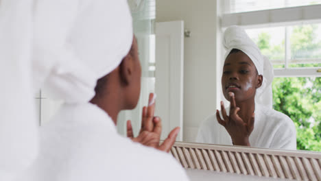 Mujer-Afroamericana-En-Bata-De-Baño-Aplicando-Crema-Facial-Mientras-Se-Mira-En-El-Espejo-En-Casa