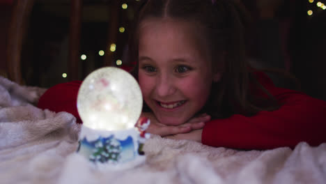 Kaukasisches-Mädchen-Lächelt-Und-Schaut-Auf-Die-Schneekugel,-Während-Sie-Zu-Weihnachten-Unter-Einer-Decke-Liegt-