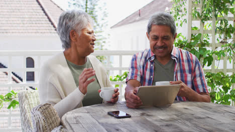 Senior-mixed-race-couple-having-coffee-using-tablet-in-garden