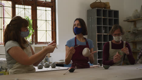 Diverse-potters-wearing-face-masks-and-aprons-painting-pots-at-pottery-studio