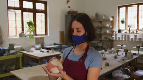 Female-caucasian-potter-wearing-face-mask-and-apron-holding-and-checking-pot-at-pottery-studio