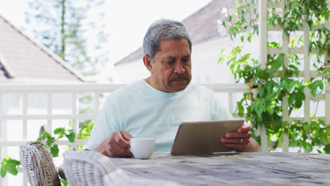 Senior-mixed-race-man-having-coffee-using-tablet-in-garden
