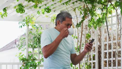 Älterer-Mann-Mit-Gemischter-Abstammung-Trinkt-Kaffee-Mit-Smartphone-Im-Garten