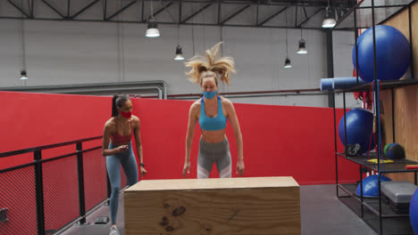 Caucasian-woman-jumping-on-box-wearing-face-mask-at-gym,-mixed-race-woman-watching