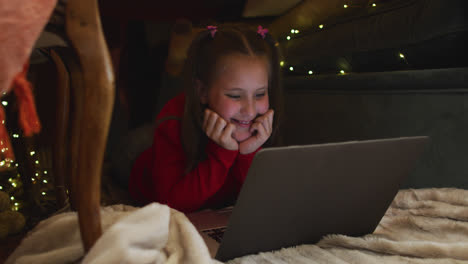 Caucasian-girl-smiling-while-using-laptop-under-blanket-fort-during-christmas-at-home