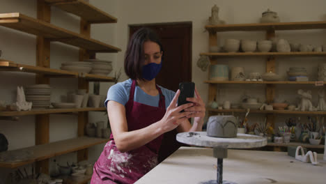 Female-caucasian-potter-wearing-face-mask-and-apron-taking-picture-of-pot-on-potters-wheel-at-potter