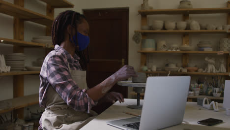Eine-Afroamerikanische-Töpferin-Mit-Gesichtsmaske-Arbeitet-An-Einem-Topf,-Während-Sie-Einen-Videochat-Auf-Dem-Laptop-Führt-