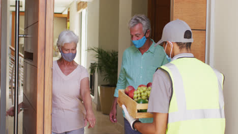 Delivery-man-delivering-groceries-to-senior-caucasian-couple-wearing-face-masks-at-home