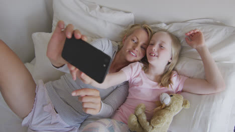 Ovear-head-view-of-Caucasian-woman-and-her-daughter-taking-selfie-on-bed