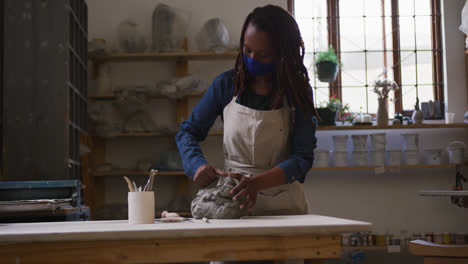 Female-potter-wearing-face-mask-and-apron-kneading-the-clay-at-pottery-studio