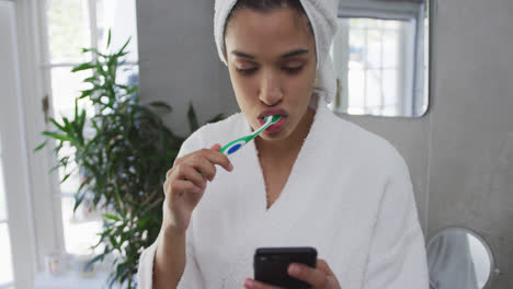 Mixed-race-woman-brushing-her-teeth-and-using-her-smartphone-in-bathroom