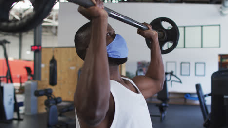 Fit-african-american-man-wearing-face-mask-exercising-using-barbell-in-the-gym