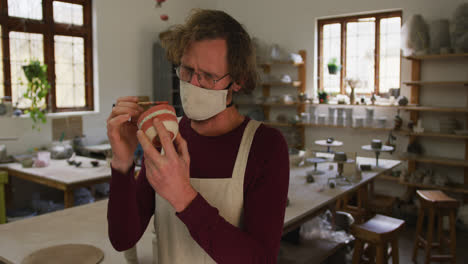 Male-caucasian-potter-wearing-face-mask-and-apron-using-brush-to-paint-pot-at-pottery-studio