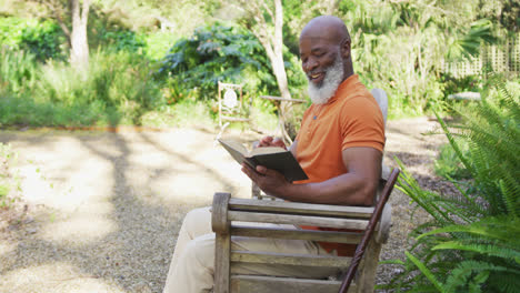 Afroamerikanischer-älterer-Mann-Lächelt,-Während-Er-Ein-Buch-Liest,-Während-Er-Auf-Einer-Bank-Im-Garten-Sitzt