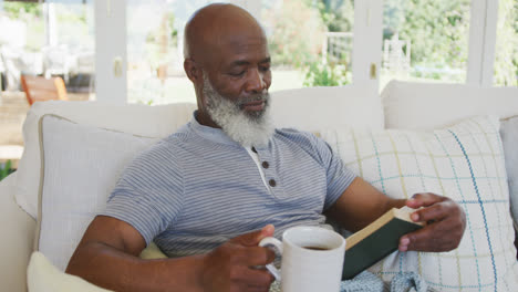 Senior-african-american-man-reading-book-and-holding-mug-of-tea