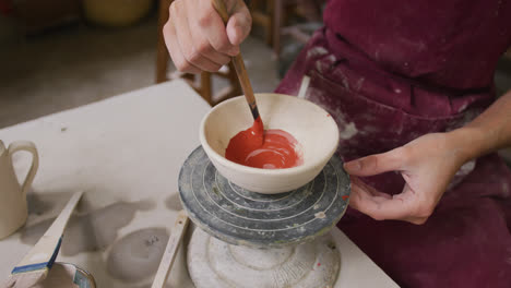 Mid-section-of-female-potter-wearing-apron-painting-pot-on-potters-wheel-at-pottery-studio