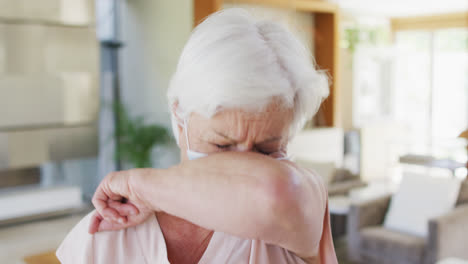 Senior-caucasian-woman-wearing-face-mask-sneezing-on-her-elbow-at-home