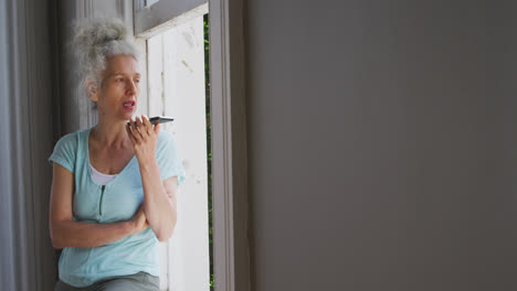 Senior-caucasian-woman-talking-on-smartphone-near-the-window-at-home