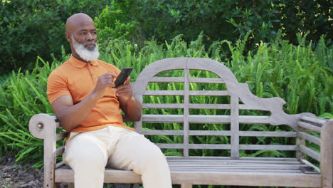 Afroamerikanischer-Senior,-Der-Sein-Smartphone-Benutzt,-Während-Er-Auf-Einer-Bank-Im-Garten-Sitzt