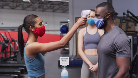 Fit-Kaukasische-Frau-Trägt-Gesichtsmaske-Misst-Temperatur-Von-Fit-Afroamerikanischen-Mann-Im-Fitnessstudio