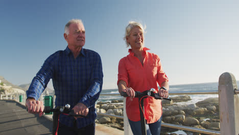 Senior-couple-walking-next-to-electronic-scooter-alongside-beach