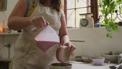 Mid-section-of-female-caucasian-potter-pouring-paint-from-a-jug-on-pot-at-pottery-studio
