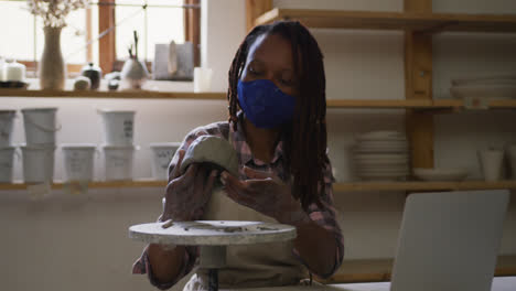 Female-african-american-potter-wearing-face-mask-working-on-pot-while-having-a-video-chat-on-laptop-