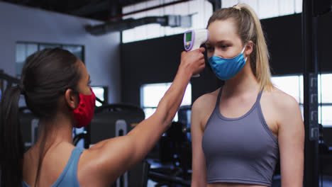 Fit-caucasian-woman-wearing-face-mask-measuring-temperature-of-fit-caucasian-woman-in-the-gym
