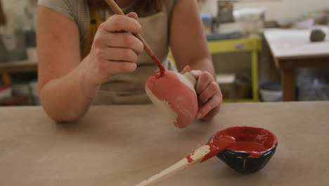 Mid-section-of-female-caucasian-potter-wearing-apron-using-brush-to-paint-pot-at-pottery-studio