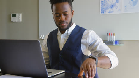 Front-view-of-young-black-businessman-sitting-with-laptop-and-talking-on-mobile-phone-in-office-4k