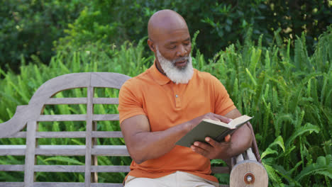 Afroamerikanischer-älterer-Mann,-Der-Ein-Buch-Liest,-Während-Er-Auf-Einer-Bank-Im-Garten-Sitzt