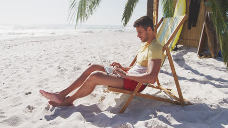 Caucasian-man-sitting-on-a-sunbed-and-using-his-laptop-on-the-beach