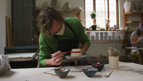Male-caucasian-potter-wearing-face-mask-and-apron-using-brush-to-paint-pot-on-potters-wheel-at-potte