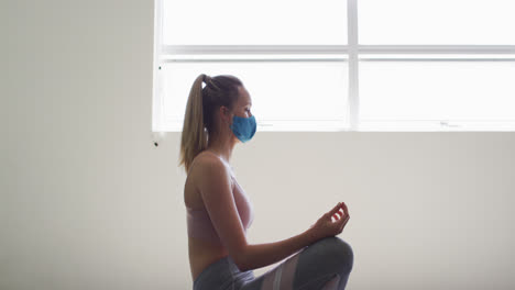 Fit-caucasian-woman-wearing-face-mask-practicing-yoga-in-yoga-studio