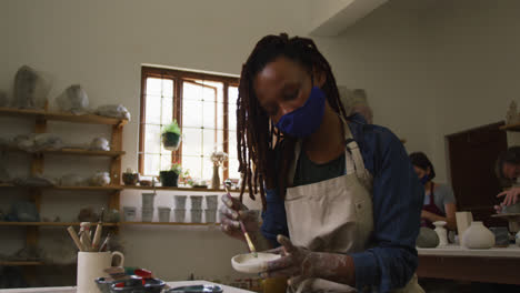 Female-potter-wearing-face-mask-and-apron-using-brush-to-paint-on-pottery-at-pottery-studio