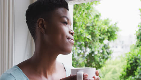 African-american-woman-holding-coffee-cup-looking-out-of-window-at-home