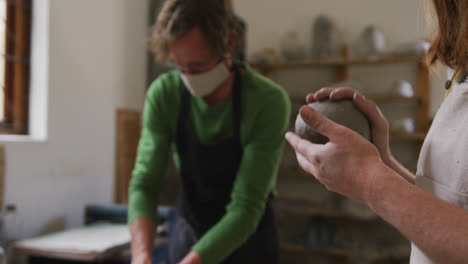 Male-and-female-potter-wearing-face-mask-and-apron-kneading-the-clay-at-pottery-studio