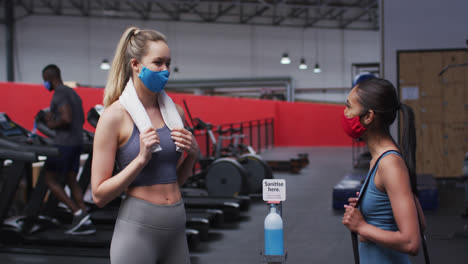 Two-fit-caucasian-women-wearing-face-mask-greeting-each-other-by-touching-elbows-in-the-gym