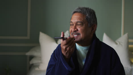Smiling-senior-mixed-race-man-sitting-on-bed-using-smartphone