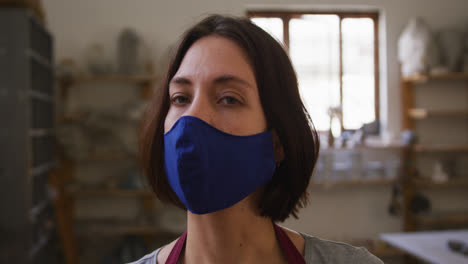 Portrait-of-female-potter-wearing-face-mask-and-apron-at-pottery-studio
