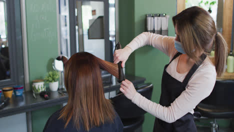 Female-hairdresser-wearing-face-mask-straightening-hair-of-female-customer-at-hair-salon